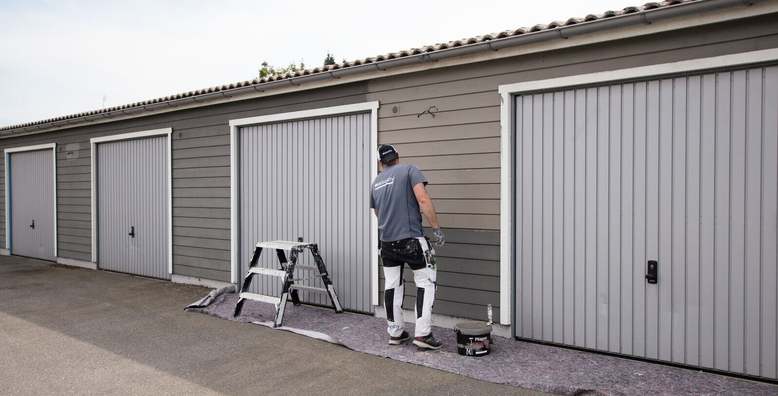 Bilden visar en målare som målar en garagefasad i en mörkgrå kulör.